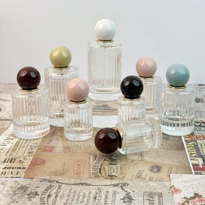 May include: A collection of clear glass perfume bottles with ribbed sides and spherical stoppers in various colors, including white, brown, black, pink, and green. The bottles have gold-colored rims.