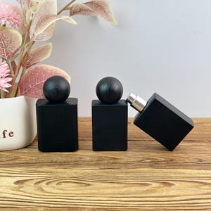 May include: Three rectangular matte black perfume bottles with spherical black wooden caps. Two bottles stand upright, while one is tilted on its side, revealing a silver metallic spray nozzle. The bottles have a smooth, opaque finish.