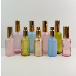 Può includere: Una collezione di flaconi spray in vetro colorato con tappi color oro. Le bottiglie sono in varie tonalità, tra cui rosa, giallo, blu e trasparente. Le bottiglie sono disposte in fila, su sfondo bianco.