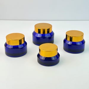 Peut inclure: Quatre pots en verre bleu cobalt avec des couvercles dorés sont disposés sur une surface blanche. Les pots sont cylindriques avec une base arrondie et un col court et large. Les couvercles sont plats et circulaires, avec une finition dorée brillante.