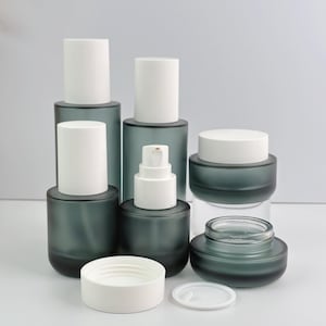 Group of frosted green glass cosmetic bottles and jars with white lids, arranged on a white surface against a white background.