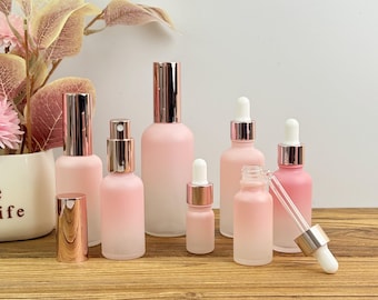 May include: A collection of pink glass bottles with metallic rose gold caps and droppers. The bottles vary in size and shape, including spray bottles and dropper bottles, and are arranged on a wooden surface. The bottles are frosted and gradient, transitioning from white to pink.
