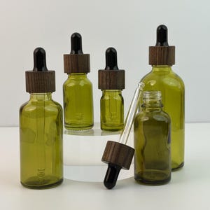 May include: An assortment of olive green glass dropper bottles with dark wood caps and black droppers. The bottles vary in size, with one bottle open and the dropper separate. The bottles are on a white surface.