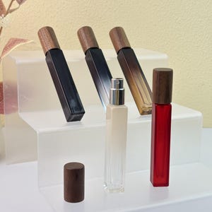 Puede incluir: Una variedad de frascos de perfume rectangulares con tapas de madera. Los frascos son de varios colores, incluyendo negro, transparente y rojo. Un frasco tiene un atomizador plateado. Los frascos se exhiben en un soporte de acrílico transparente.