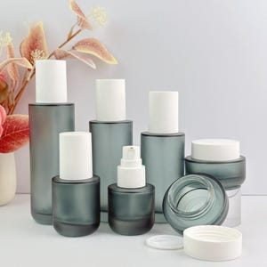 Arrangement of frosted green glass skincare containers including pump bottles and cream jars, with floral decorations in the background. One cream jar is open showing the inside.
