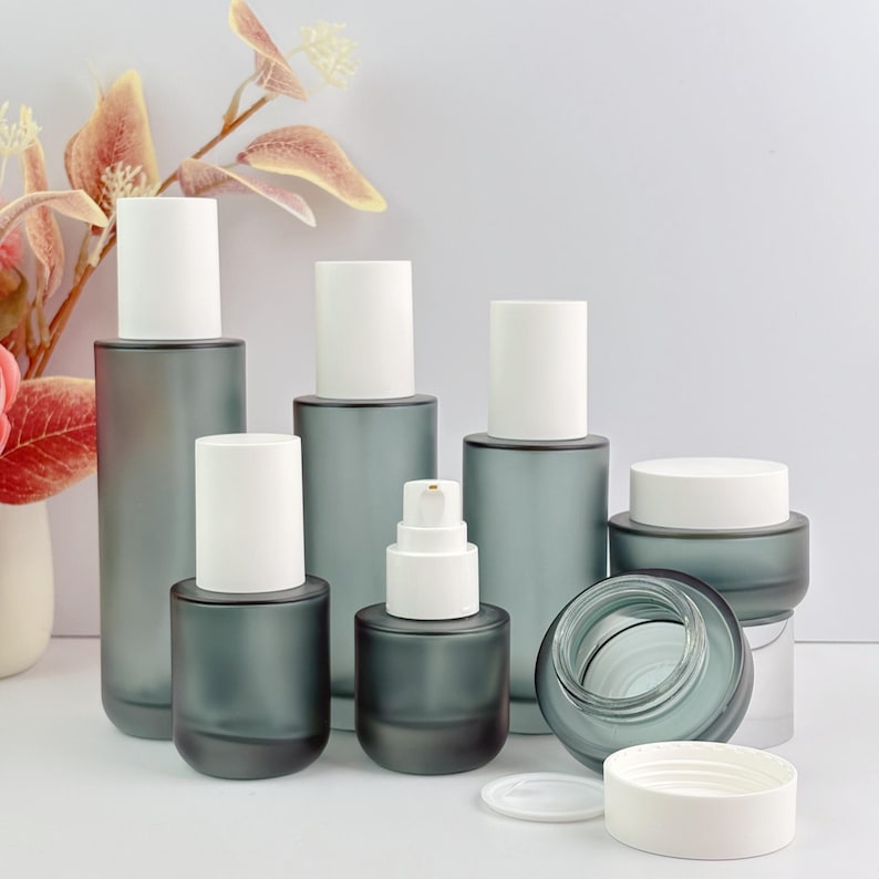 Arrangement of frosted green glass skincare containers including pump bottles and cream jars, with floral decorations in the background. One cream jar is open showing the inside.