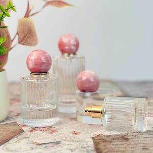 Op de afbeelding: Transparante glazen parfumflesjes met roze, marmerachtige, bolvormige stoppen. De flesjes hebben een geribbeld ontwerp en goudkleurige sproeiers. De achtergrond omvat bloemig patroonpapier en decoratieve elementen.