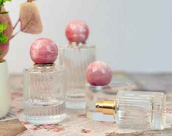 May include: Clear glass perfume bottles with pink marble-like spherical stoppers. The bottles have a ribbed design and gold-colored spray nozzles. The background includes floral patterned paper and decorative elements.