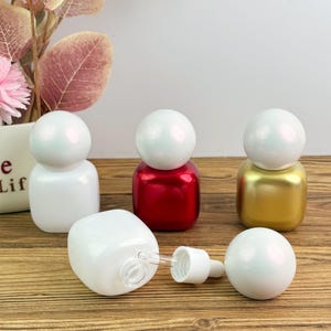 May include: A collection of decorative glass dropper bottles in white, red, and gold. The bottles have a rounded top and a square base. One bottle is lying on its side with the dropper exposed. The background includes a vase with flowers.