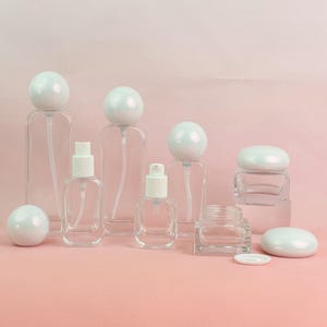 Puede incluir: Colección de botellas y tarros de vidrio transparente con tapones y tapas blancos de forma esférica y ovalada. Las botellas incluyen pulverizadores y un tarro, todos dispuestos sobre una superficie rosa. Las botellas varían en tamaño y forma.