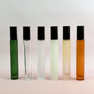 Könnte beinhalten: Sechs Roll-on-Parfümflaschen mit schwarzen Kappen. Die Flaschen sind in verschiedenen Farben erhältlich, darunter Grün, Klar, mattiertes Weiß, Hellgrün, Gelb und Bernstein. Die Flaschen sind in einer Reihe vor einem schlichten Hintergrund angeordnet.