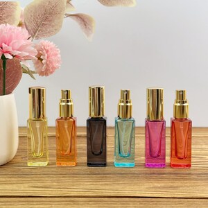 May include: Six rectangular glass spray bottles with gold-colored caps arranged in a row. The bottles are colored yellow, orange, black, light blue, pink, and red. Each bottle features a clear plastic spray nozzle and a transparent internal tube.