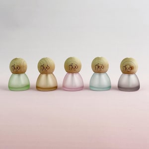 Può includere: Cinque piccole bottiglie decorative in vetro in fila, ciascuna con un tappo di legno. Le bottiglie sono in tonalità di verde, beige, rosa, blu e grigio. Ogni tappo di legno ha un piccolo disegno inciso. Le bottiglie hanno una forma arrotondata.