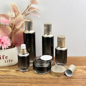 Könnte beinhalten: Eine Sammlung von dunkelgrauen Glaskosmetikbehältern mit goldfarbenen, gerippten Verschlüssen. Das Set enthält zwei hohe Flaschen, eine kleinere Pumpflasche und einen breiten Tiegel mit weißem Innendeckel. Die Behälter haben ein Farbverlauf-Finish.