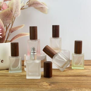 May include: A collection of square, frosted glass perfume bottles with dark brown wooden caps. Some bottles are standing upright, one is lying on its side, and one has its wooden cap removed to reveal a silver spray nozzle mechanism.
