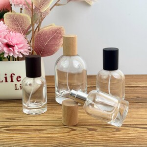 May include: Clear glass perfume bottles with wooden and black caps are arranged on a wooden surface. The bottles vary in size and shape, with a small white flower pot and pink flowers in the background. The bottles are empty and ready to be filled.
