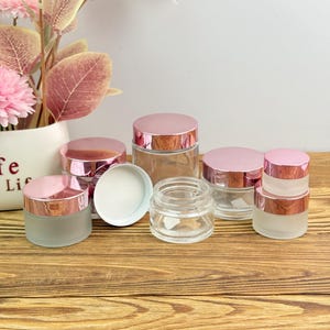 Puede incluir: Una colección de frascos de vidrio transparente y esmerilado con tapas de oro rosa. Los frascos varían en tamaño y están dispuestos sobre una superficie de madera. Algunos frascos están abiertos, revelando un interior blanco.