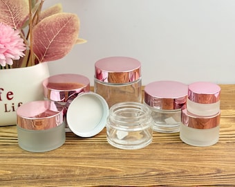 May include: A collection of clear and frosted glass jars with rose gold lids. The jars vary in size and are arranged on a wooden surface. Some jars are open, revealing a white interior.
