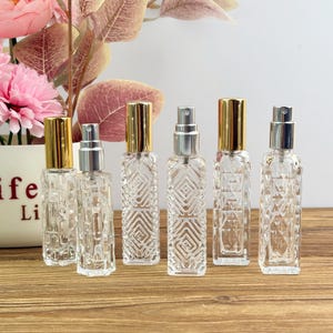 Puede incluir: Seis frascos de perfume de vidrio transparente con pulverizadores dorados y plateados están dispuestos sobre una superficie de madera. Los frascos tienen forma rectangular con un diseño geométrico texturizado. Un jarrón con flores rosas está en el fondo.