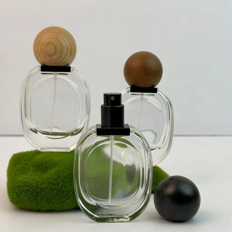 Three identical white transparent glass cosmetic containers, each topped with a differently colored spherical wooden cap—black, brown, and light wood—are arranged in staggered inverted triangles. The light wood glass spray bottle rests atop a moss-covered stone on the left, while the perfume bottle with its black spray nozzle sits forward, its matching black cap placed on the moss-covered stone to the right.
