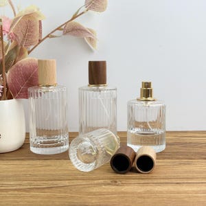 Op de afbeelding: Heldere glazen parfumflessen met houten en goudkleurige sproeikoppen. De flessen hebben een geribbeld ontwerp en worden weergegeven op een houten oppervlak. Twee houten doppen zijn ook zichtbaar. Een decoratief bloemstuk staat op de achtergrond.