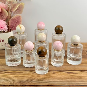 Pode incluir: Uma coleção de frascos de perfume de vidro transparente com tampas decorativas de pedra em várias cores, incluindo rosa, castanho e branco. Os frascos têm um design canelado e colares dourados. Os frascos estão dispostos sobre uma superfície de madeira.