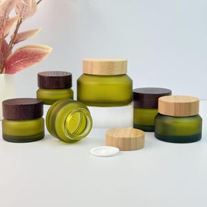 Op de afbeelding: Een assortiment olijfgroene glazen potten met houten deksels. Sommige deksels zijn lichtgekleurd hout, andere zijn donkerbruin. Eén pot is open en onthult de binnenkant. De potten zijn van verschillende grootte en staan op een wit oppervlak.