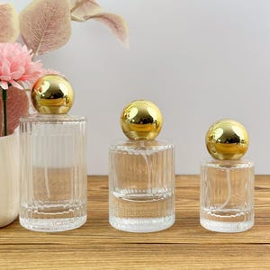 Peut inclure: Trois flacons de parfum en verre transparent de hauteurs différentes, chacun présentant une texture côtelée verticale sur le corps et un bouchon sphérique doré lisse et réfléchissant. Les flacons sont vides et incluent un tube pulvérisateur.