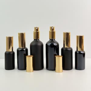 Puede incluir: Un surtido de botellas de spray y botellas pequeñas de vidrio negro con tapas doradas. Las botellas varían en tamaño y forma, algunas tienen un acabado mate y otras brillante. Las tapas son cilíndricas y brillantes.