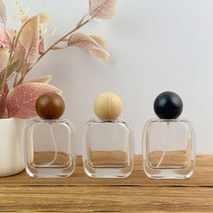 May include: Three square glass perfume bottles with rounded corners and spherical caps. The left bottle has a dark brown wooden cap, the center bottle has a light natural wood cap, and the right bottle has a solid black spherical cap.