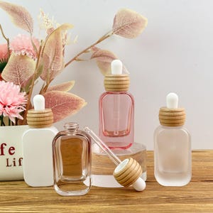 May include: Four rectangular glass dropper bottles with rounded corners and wooden collars. One bottle is opaque white, one is clear, one is translucent pink, and one is frosted. Each features a white rubber dropper top and a glass pipette.