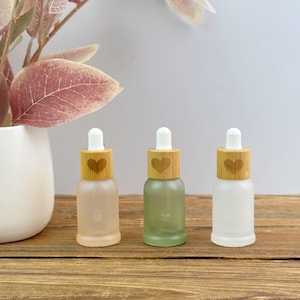 May include: Three small glass dropper bottles with frosted finishes in clear, light green, and white. Each bottle features a light-colored wooden collar engraved with a heart icon and a white rubber dropper bulb top for dispensing liquid contents.