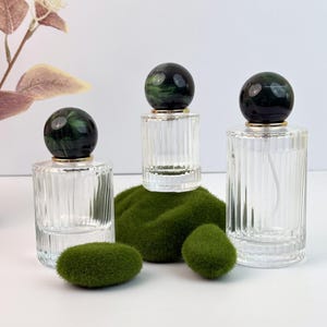 Puede incluir: Tres frascos de perfume de vidrio transparente con lados acanalados y tapones esféricos de mármol negro y verde oscuro. Los frascos están colocados sobre un lecho de musgo verde, con dos piezas de musgo adicionales delante.
