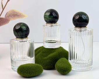 May include: Three clear glass perfume bottles with ribbed sides and dark green and black marble-like spherical stoppers. The bottles are arranged on a bed of green moss, with two additional moss pieces in front.