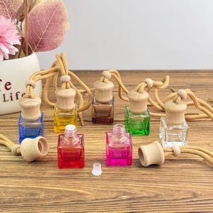 May include: A selection of small, square glass bottles in various colours, each with a wooden cap and a beige cord for hanging. The bottles are filled with coloured liquid and are arranged on a wooden surface.
