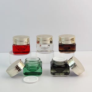 Peut inclure: Cinq pots cosmétiques carrés en verre avec des couvercles vissables en métal doré. Trois pots sont fermés et disposés à l'arrière, en verre rouge, transparent et ambré. Deux pots sont à l'avant, un vert et un gris foncé, sans couvercle.