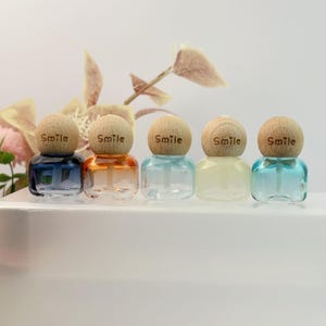 Può includere: Cinque piccole bottiglie di vetro trasparente con tappi di legno, ognuna con la scritta "Smile". Le bottiglie contengono liquidi colorati: blu, arancione, trasparente, giallo e turchese. Le bottiglie sono disposte in fila.