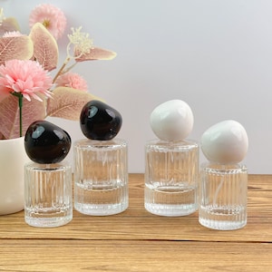 Op de afbeelding: Vier doorzichtige glazen parfumflesjes met verticale geribbelde texturen op de basis. Twee flesjes hebben gladde, ronde zwarte doppen, terwijl de andere twee flesjes gladde, ronde witte doppen hebben. De flesjes staan op een rij.