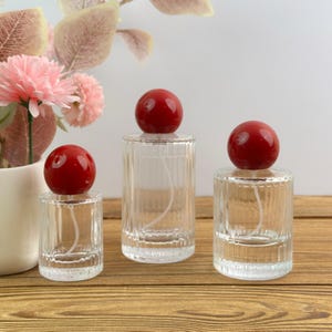 Peut inclure: Trois flacons de parfum en verre transparent de hauteurs différentes, chacun présentant une texture côtelée verticale sur le corps et un bouchon sphérique rouge brillant et lisse. Les flacons sont transparents, révélant un tube pulvérisateur.
