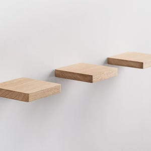 May include: Three square, floating shelves made of light-colored wood are mounted on a white wall. The shelves are evenly spaced and appear to be made of solid wood, showcasing the natural grain.