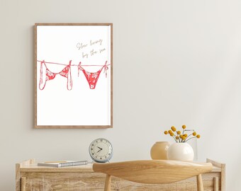 May include: A framed print featuring a red bikini with a white floral pattern hanging on a clothesline. The words "Slow living by the sea" are written in cursive. The print is displayed on a wooden desk with a clock and vases.