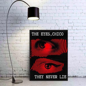 May include: Black framed art print with two red spiral eye designs on a black background. The text "THE EYES, CHICO" and "THEY NEVER LIE" are printed in white. The art is displayed next to a black floor lamp.