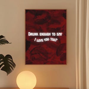 May include: A framed print with a red background of lipstick marks. The white text reads "Drunk enough to say I love you too?" The print is in a wooden frame and is displayed on a wall.