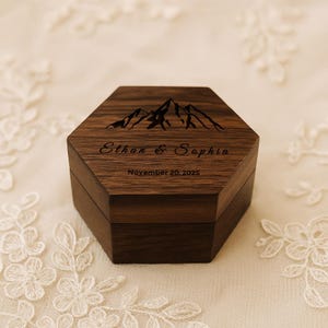May include: A dark brown, hexagonal wooden ring box. The lid features a mountain range design and the names "Ethan & Sophia" with the date "November 20, 2025" engraved below. The box is set against a soft, white lace background.