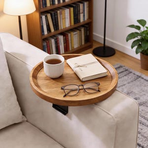 May include: A round wooden sofa arm tray with a cup of tea, a notebook, and eyeglasses. The tray is attached to the arm of a light-colored sofa. A bookshelf and lamp are in the background.