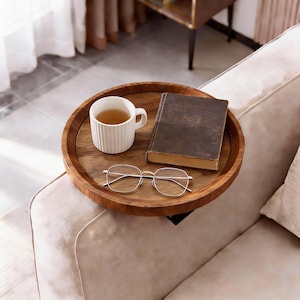 Könnte beinhalten: Ein runder Holztablett mit einer weißen Tasse, einem alten Buch und einer Brille mit silbernem Rahmen. Das Tablett steht auf einer beigen Sofalehne und schafft eine entspannte Atmosphäre. Die Gegenstände deuten auf einen ruhigen Moment mit einem Buch und Tee hin.