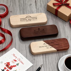 May include: Three wooden pen cases, two-toned and solid, with engraved text. One case is light wood, another is dark brown, and the third is a combination of both. A Christmas card and gift box with a red ribbon are also visible.