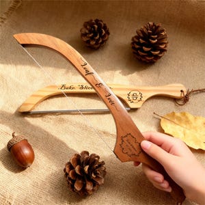 May include: A wooden bread slicer with a curved handle and a stainless steel wire. The handle is engraved with the words "Loaf & Love" and a decorative design. A second bread slicer is in the background. The image includes pine cones and an acorn.