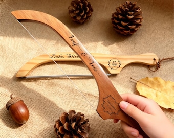 May include: A wooden bread slicer with a curved handle and a stainless steel wire. The handle is engraved with the words "Loaf & Love" and a decorative design. A second bread slicer is in the background. The image includes pine cones and an acorn.