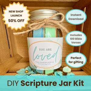 Könnte beinhalten: Ein DIY-Schriftglas-Kit in einem Einmachglas mit silbernem Deckel, gefüllt mit bunten Bändern. Das Glas hat ein Etikett mit der Aufschrift "YOU ARE loved" und enthält 100 Bibelverse. Das Kit ist mit Bindfaden gebunden.